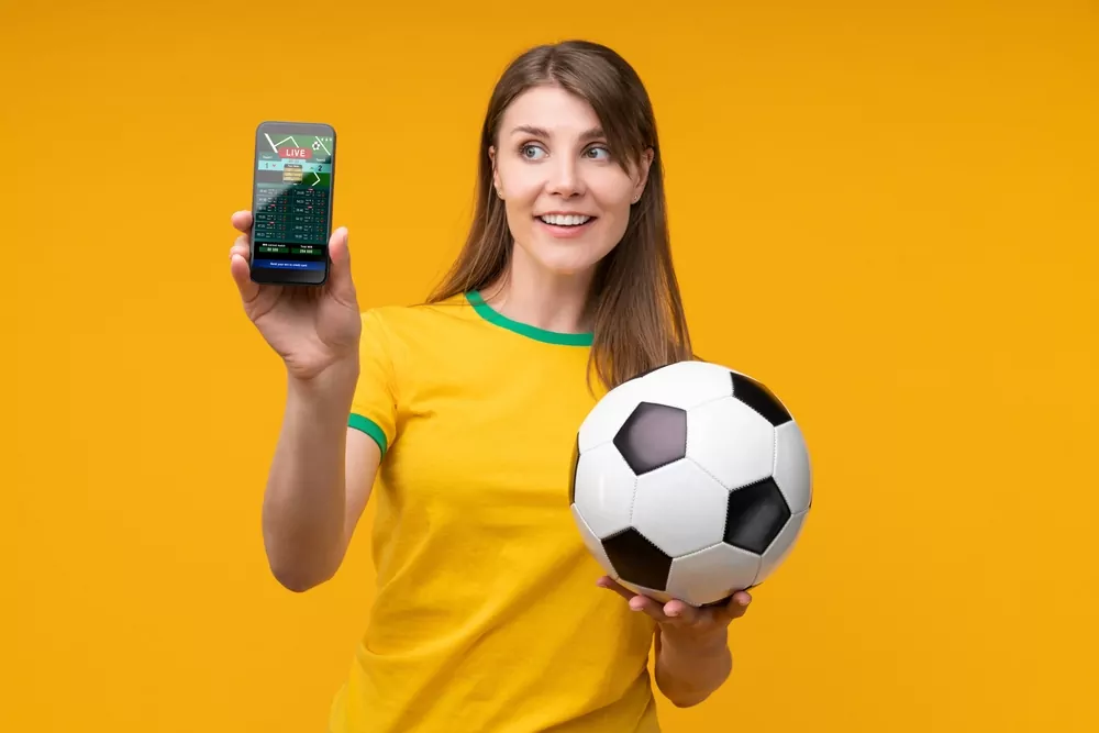 Fußballfan hält ein Smartphone mit Sportwetten App und einen Ball in der Hand