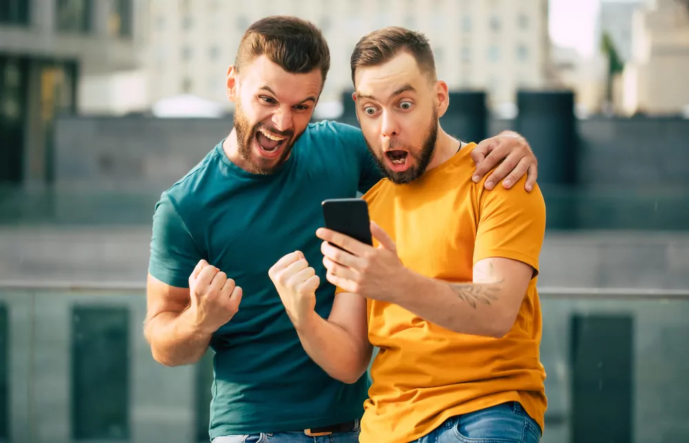 Zwei Fußballfans jubeln über einen Treffer in der Sportwetten App auf dem Smartphone