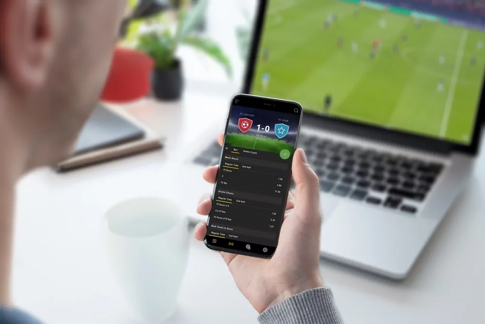 Smartphone mit Wett App und laufendem Fußballspiel auf dem Laptop im Hintergrund