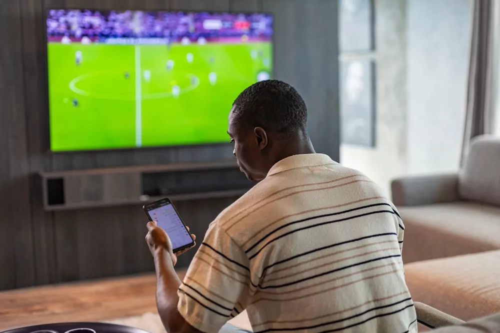 Mann platziert in Ruhe eine Wette in der App, während im Fernsehen ein Fußballspiel läuft